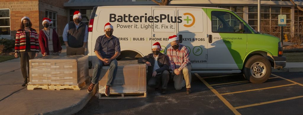 Batteries Plus employees in Christmas hats sitting on pallets of toys in front of a Batteries Plus van
