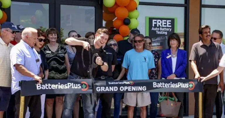 News 3 A man cuts a ribbon at the grand opening of a Batteries Plus store, surrounded by a group of people. The storefront has a "FREE Auto Battery Testing" sign and balloons.