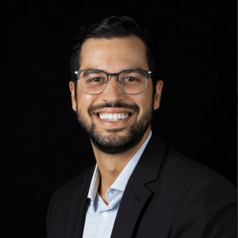 Batteries Plus VP Empowers Dreams Through Franchising 4 A man with glasses and a beard is smiling while wearing a suit jacket over a light blue shirt, posing against a black background.